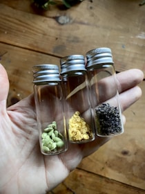Three small glass vials with silver caps are held in a hand. Each vial contains different types of seeds or spices: green cardamom pods, yellow mustard seeds, and dark peppercorns. The wooden surface in the background adds a rustic feel.