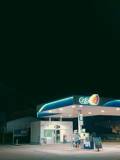 A gas station is illuminated at night, showcasing bright artificial lights. The station has a modern canopy with a prominent logo on display. There is a shop attached to the gas station, and the area is quiet with no visible vehicles or people.