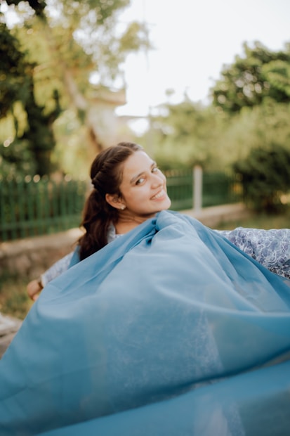 A person is joyfully spinning outdoors, with a flowing blue fabric creating a dynamic and lively scene. The background is a lush green park with trees and a fence, suggesting a sunny day.