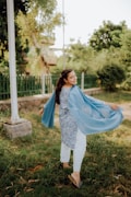 A woman in traditional attire is twirling joyfully in a grassy park. Her blue scarf flutters behind her as she turns. The background features lush greenery and a fence, with soft, natural lighting creating a serene atmosphere.