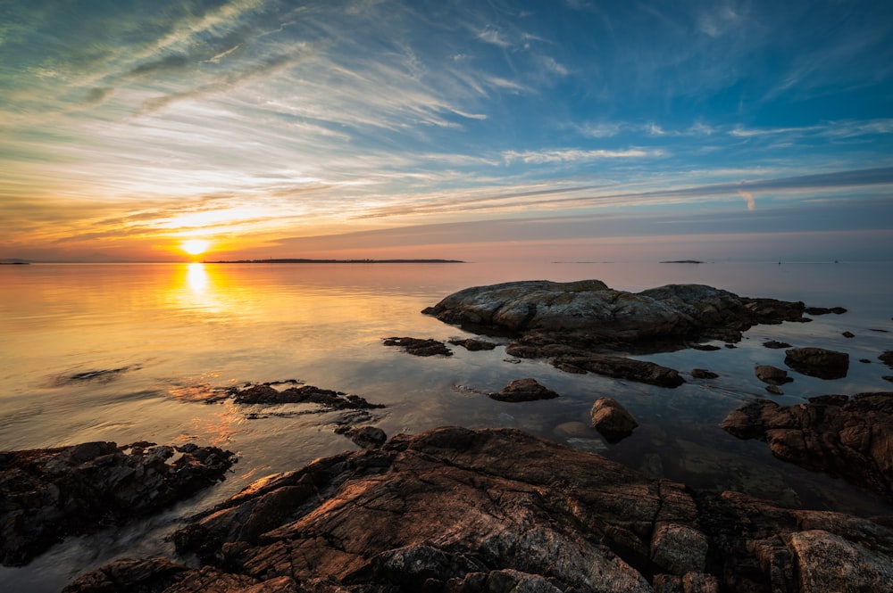 the sun is setting over the ocean with rocks in the foreground