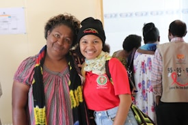 Two people are posing together, with one person wearing a red shirt with a logo and a black beanie, while the other wears a colorful dress with a scarf. In the background, more people are visible, some wearing shirts with health-related messages.