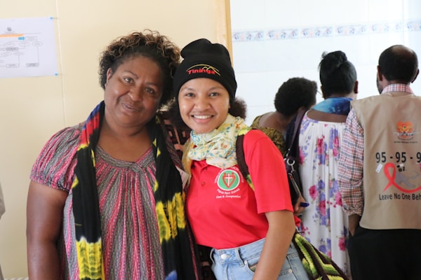 Two people are posing together, with one person wearing a red shirt with a logo and a black beanie, while the other wears a colorful dress with a scarf. In the background, more people are visible, some wearing shirts with health-related messages.