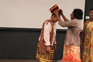 Several people are interacting, with one person wearing traditional attire while another helps adjust a vibrant, multicolored hat on their head. The group appears to be indoors, with a simple wall background and wooden flooring.