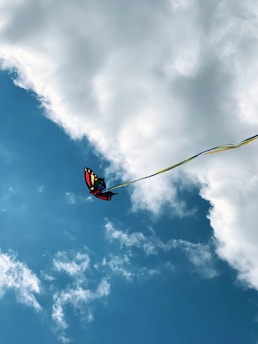 A colorful butterfly-shaped kite flying against a bright blue sky with large, fluffy white clouds. The kite is attached to a yellow and blue string that extends out of the frame.