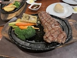 A sizzling dish featuring a grilled steak with visible grill marks, accompanied by vibrant green broccoli, orange baby carrots, and a piece of yellow corn on a black sizzling platter. A piece of bone marrow with herbs and mashed potatoes is also included. In the background, there is a small dish of sauce and a soft bread roll on a white plate.