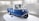 A small blue and white vintage vehicle with three wheels. The vehicle has a compact design and is placed on a circular platform inside a well-lit exhibition space with vertical white slats on the background walls. The vehicle has a unique front design with round headlights.