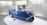 A small blue and white vintage vehicle with three wheels. The vehicle has a compact design and is placed on a circular platform inside a well-lit exhibition space with vertical white slats on the background walls. The vehicle has a unique front design with round headlights.
