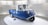 A small blue and white vintage vehicle with three wheels. The vehicle has a compact design and is placed on a circular platform inside a well-lit exhibition space with vertical white slats on the background walls. The vehicle has a unique front design with round headlights.