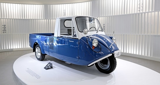A small blue and white vintage vehicle with three wheels. The vehicle has a compact design and is placed on a circular platform inside a well-lit exhibition space with vertical white slats on the background walls. The vehicle has a unique front design with round headlights.