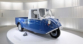 A small blue and white vintage vehicle with three wheels. The vehicle has a compact design and is placed on a circular platform inside a well-lit exhibition space with vertical white slats on the background walls. The vehicle has a unique front design with round headlights.
