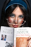 A person with long dark hair and a turquoise headband is holding a book by Orhan Pamuk titled 'Kirmizi Sacli Kadin'. The person is wearing glasses with a reflective surface and has a focused expression. The book cover has an image of Orhan Pamuk and a piece of art, along with text in Turkish.