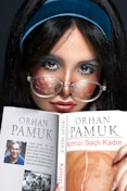 A person with long dark hair and a turquoise headband is holding a book by Orhan Pamuk titled 'Kirmizi Sacli Kadin'. The person is wearing glasses with a reflective surface and has a focused expression. The book cover has an image of Orhan Pamuk and a piece of art, along with text in Turkish.