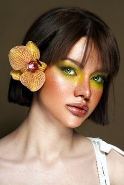 A woman with striking green eyeshadow and pronounced freckles gazes forward. She has short brown hair, accented with a yellow orchid. Her expression is calm and confident, and she wears a white top.