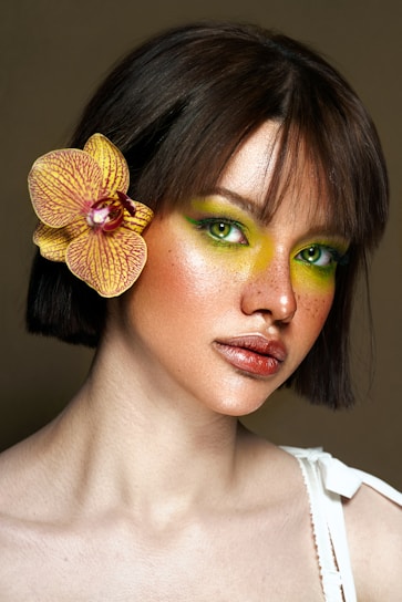 A woman with striking green eyeshadow and pronounced freckles gazes forward. She has short brown hair, accented with a yellow orchid. Her expression is calm and confident, and she wears a white top.