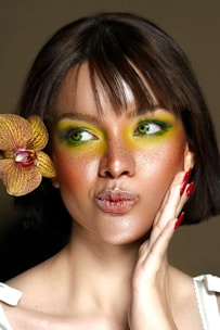 A person with short brown hair and vibrant green eye makeup holds an orchid flower close to their face. The expression is playful, with lips slightly puckered and a hand resting on the cheek, highlighting neatly manicured nails painted in red. The lighting emphasizes the smooth skin and freckles, creating a striking portrait against a neutral background.