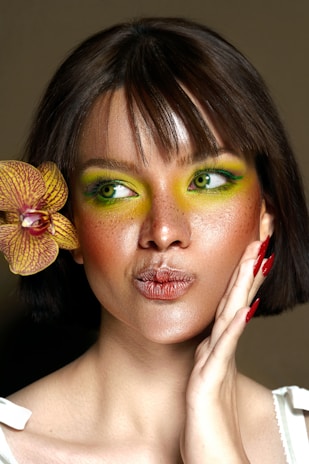 A person with short brown hair and vibrant green eye makeup holds an orchid flower close to their face. The expression is playful, with lips slightly puckered and a hand resting on the cheek, highlighting neatly manicured nails painted in red. The lighting emphasizes the smooth skin and freckles, creating a striking portrait against a neutral background.