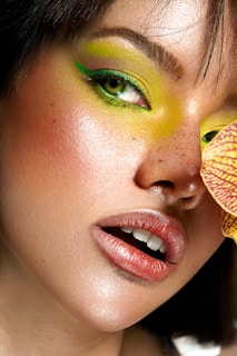 A close-up of a person's face highlighting artistic makeup. The eyes are accentuated with vibrant green eyeshadow extending to the eyebrows. The skin is smooth with freckles visible, and lips are glossed in a muted pink shade. A piece of brightly patterned yellow and red fabric is partially visible.