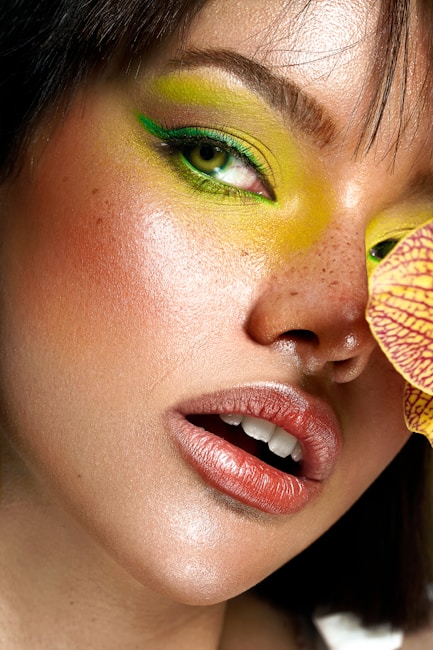 A close-up of a person's face highlighting artistic makeup. The eyes are accentuated with vibrant green eyeshadow extending to the eyebrows. The skin is smooth with freckles visible, and lips are glossed in a muted pink shade. A piece of brightly patterned yellow and red fabric is partially visible.