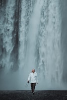 A person in a white jacket walks towards the camera holding a camera, with a massive waterfall cascading down behind them. The environment appears misty and the ground is dark and wet, creating a dramatic and serene scene.
