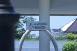 A wheelchair-accessible sign is mounted on a curved metal frame. The background features a partly sunny outdoor scene with trees, a structure with a sloped roof, and a clear sky.