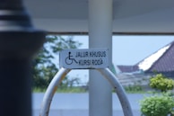 A wheelchair-accessible sign is mounted on a curved metal frame. The background features a partly sunny outdoor scene with trees, a structure with a sloped roof, and a clear sky.