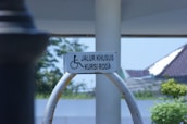A wheelchair-accessible sign is mounted on a curved metal frame. The background features a partly sunny outdoor scene with trees, a structure with a sloped roof, and a clear sky.