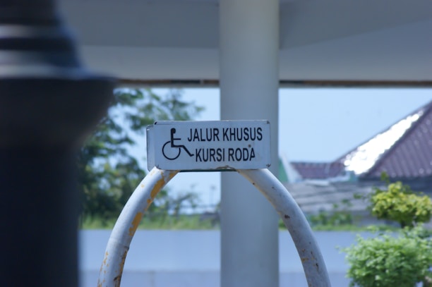 A wheelchair-accessible sign is mounted on a curved metal frame. The background features a partly sunny outdoor scene with trees, a structure with a sloped roof, and a clear sky.