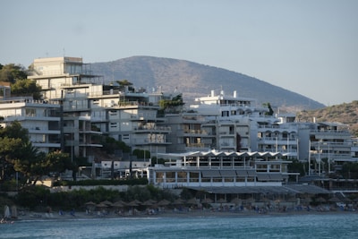 A cluster of modern, multi-story buildings along a coastal area with mountains in the background. The architecture is contemporary with flat roofs and glass balconies. Below, there is a beach lined with umbrellas and people enjoying the seaside.