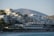 A cluster of modern, multi-story buildings along a coastal area with mountains in the background. The architecture is contemporary with flat roofs and glass balconies. Below, there is a beach lined with umbrellas and people enjoying the seaside.