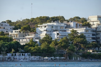 A picturesque beach scene featuring several modern white buildings nestled among lush green trees on a hillside. In the foreground, the calm blue sea extends to the sandy shore, where sun loungers and umbrellas are positioned. People are seen enjoying the water, contributing to a lively yet relaxed atmosphere.