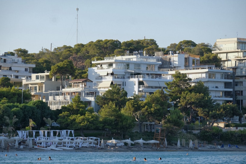 A picturesque beach scene featuring several modern white buildings nestled among lush green trees on a hillside. In the foreground, the calm blue sea extends to the sandy shore, where sun loungers and umbrellas are positioned. People are seen enjoying the water, contributing to a lively yet relaxed atmosphere.