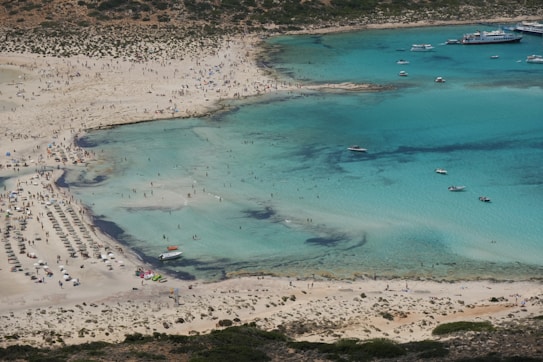 A vast sandy beach is filled with sun umbrellas and people sunbathing, while the clear turquoise water hosts a few boats and swimmers. The shoreline is dotted with small bushes and the bay is surrounded by rocky terrain. A large ferry is moored at a dock on the far side of the bay.