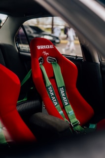 A red racing seat with Takata brand green harnesses is installed in the interior of a car. The seat features the Bride logo and is designed for high-performance driving. The car's interior is mostly dark, providing a contrast to the bright seat and harness. The outside environment is blurred, indicating the focus on the seat.