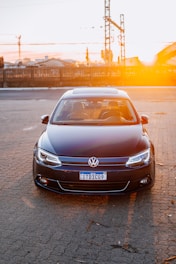 A dark-colored Volkswagen car is parked on a paved surface with a setting sun in the background creating a warm glow. The car is facing forward, displaying its front features and a Brazilian license plate. Industrial structures and a wooden fence are visible in the distance.