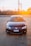 A dark-colored Volkswagen car is parked on a paved surface with a setting sun in the background creating a warm glow. The car is facing forward, displaying its front features and a Brazilian license plate. Industrial structures and a wooden fence are visible in the distance.