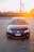 A dark-colored Volkswagen car is parked on a paved surface with a setting sun in the background creating a warm glow. The car is facing forward, displaying its front features and a Brazilian license plate. Industrial structures and a wooden fence are visible in the distance.