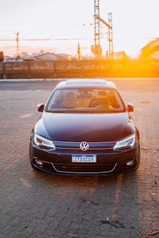 A dark-colored Volkswagen car is parked on a paved surface with a setting sun in the background creating a warm glow. The car is facing forward, displaying its front features and a Brazilian license plate. Industrial structures and a wooden fence are visible in the distance.