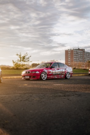 A customized red car with bold graphics and large wheels parked on a paved area. In the background, there is a large brick building, some trees, and a fence. The setting sun casts a warm glow over the scene, creating a dramatic light effect.