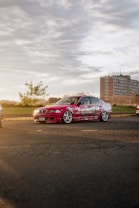 A customized red car with bold graphics and large wheels parked on a paved area. In the background, there is a large brick building, some trees, and a fence. The setting sun casts a warm glow over the scene, creating a dramatic light effect.