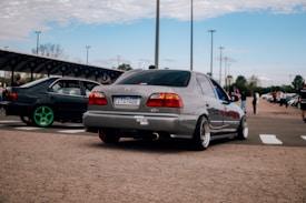 A parking lot with two modified cars, one in the forefront with a silver body and aftermarket wheels and another in the background with bright green wheels. The setting is outdoors with a clear blue sky, a few scattered clouds, and several people casually walking around in the background. Light poles and a shaded structure can be seen lining the parking area.