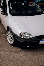 A close-up view of a parked white car with a clean and shiny exterior. The focus is on the front section of the car, showcasing its headlight, grille, and polished alloy wheel. The car is parked on an asphalt surface, with part of its registration plate visible.