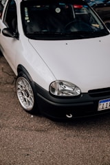 A close-up view of a parked white car with a clean and shiny exterior. The focus is on the front section of the car, showcasing its headlight, grille, and polished alloy wheel. The car is parked on an asphalt surface, with part of its registration plate visible.