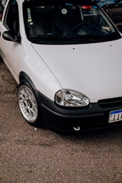 A close-up view of a parked white car with a clean and shiny exterior. The focus is on the front section of the car, showcasing its headlight, grille, and polished alloy wheel. The car is parked on an asphalt surface, with part of its registration plate visible.