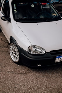 A close-up view of a parked white car with a clean and shiny exterior. The focus is on the front section of the car, showcasing its headlight, grille, and polished alloy wheel. The car is parked on an asphalt surface, with part of its registration plate visible.