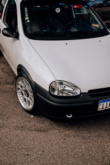 A close-up view of a parked white car with a clean and shiny exterior. The focus is on the front section of the car, showcasing its headlight, grille, and polished alloy wheel. The car is parked on an asphalt surface, with part of its registration plate visible.