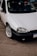 A close-up view of a parked white car with a clean and shiny exterior. The focus is on the front section of the car, showcasing its headlight, grille, and polished alloy wheel. The car is parked on an asphalt surface, with part of its registration plate visible.