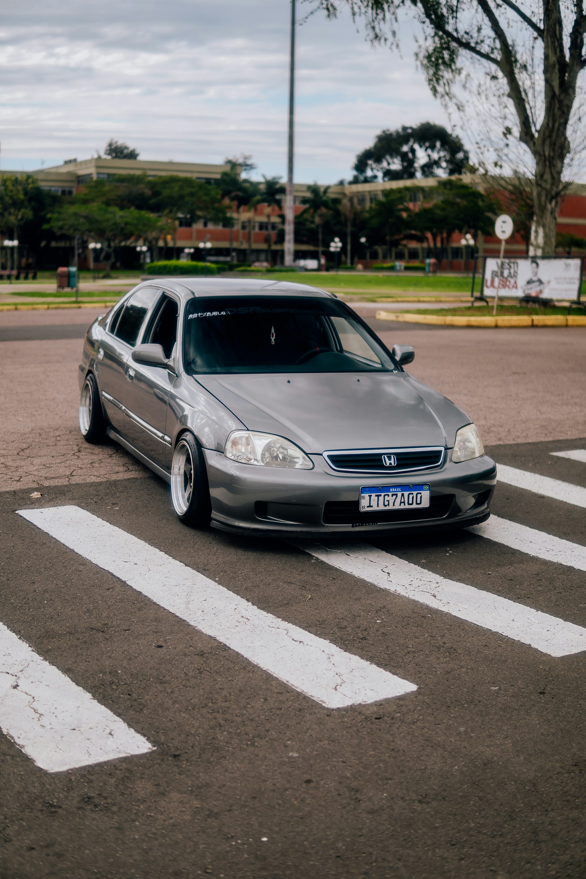 A gray Honda sedan is parked on a pedestrian crosswalk. The setting appears to be an urban area with buildings and trees in the background. The car has custom rims and lowered suspension. A license plate reads '1TG7AGO'. The sky is overcast, indicating cloudy weather.
