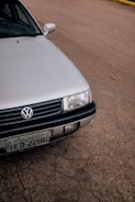A close-up view of the front portion of a silver Volkswagen car, parked on a paved surface. The vehicle's grille, headlight, and license plate are visible. The background consists of a textured asphalt road, with a small section of a yellow curb in the distance.