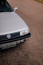 A close-up view of the front portion of a silver Volkswagen car, parked on a paved surface. The vehicle's grille, headlight, and license plate are visible. The background consists of a textured asphalt road, with a small section of a yellow curb in the distance.
